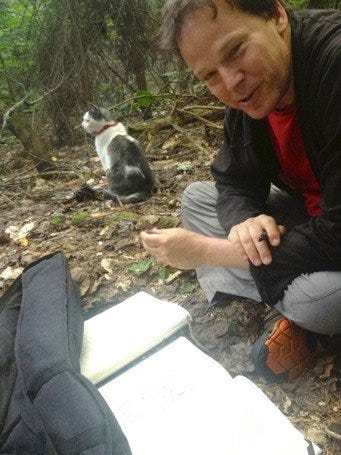 David Graeber sitting cross-legged in forest with a book in frnt of him and a cat to one side