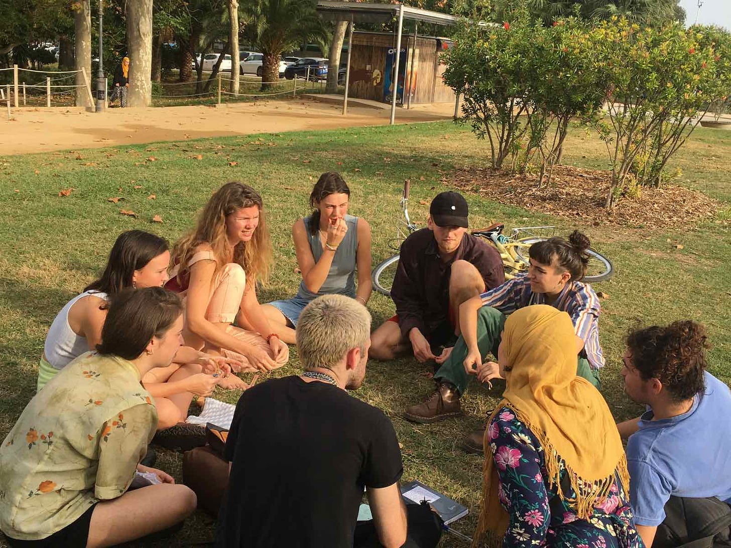 A group of around 10 students sit in a circle on some grass in discussion on a sunny day