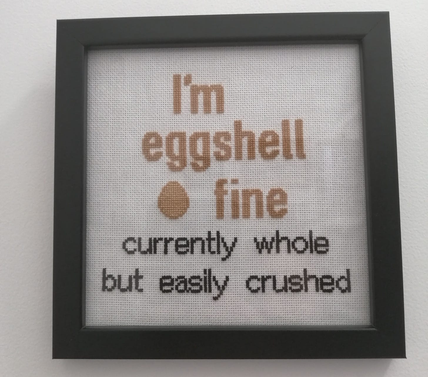 Cross stitch in a frame with eggshell coloured text that reads "I'm eggshell fine" with a picture of an egg then black text that reads "currently whole but easily crushed"