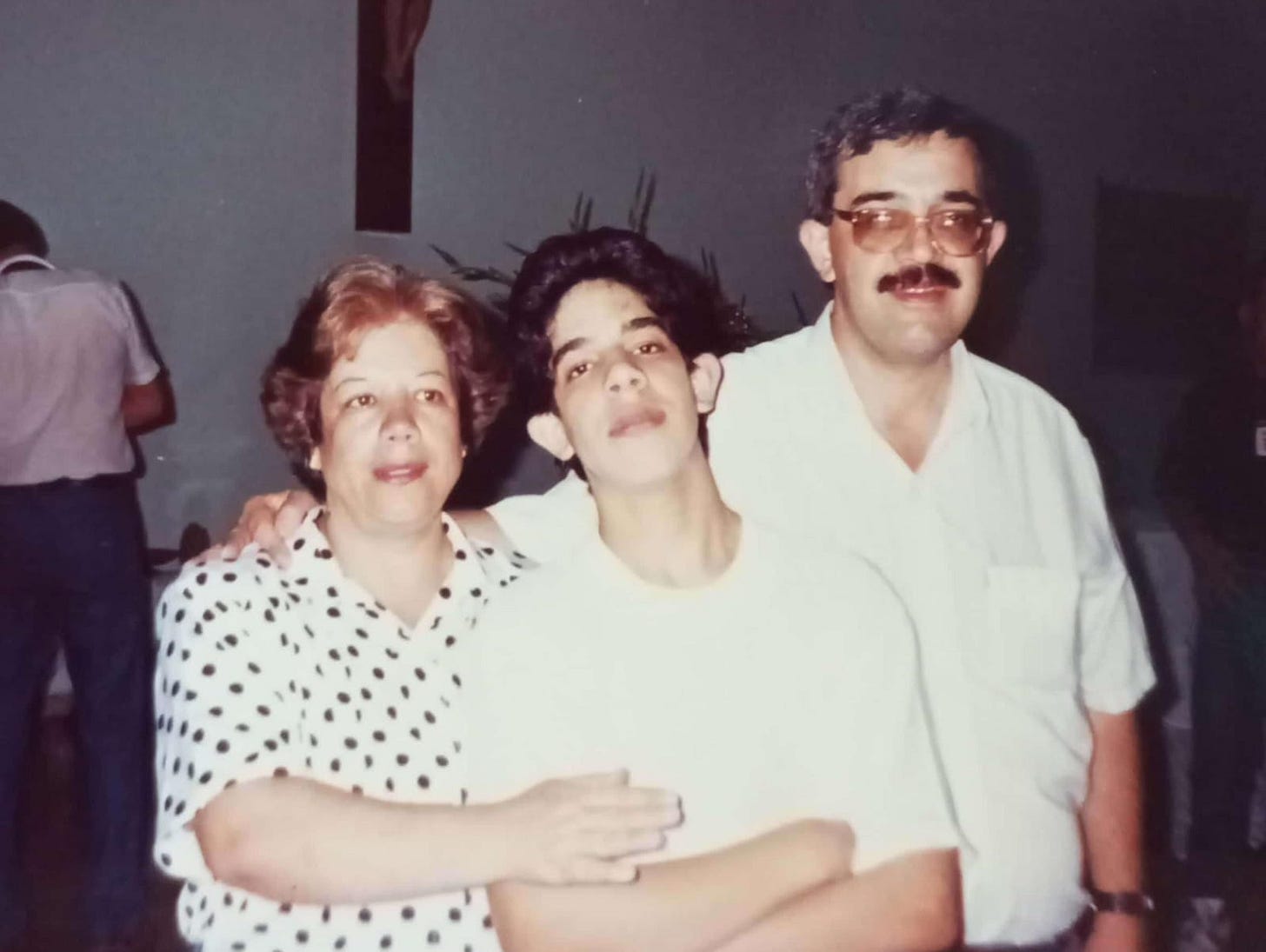 Imagem mostra um casal em volta do filho adolescente, também fotografados em plano americano. Ela tem cabelos curtos e ondulados, castanhos, com a franja mais clara; veste camisa social branca com bolinhas pretas; o jovem tem cabelos pretos curtos e usa camiseta branca de manga curta; o homem usa óculos e bigode, tem cabelos pretos com alguns fios brancos e usa camisa social branca de manga curta.