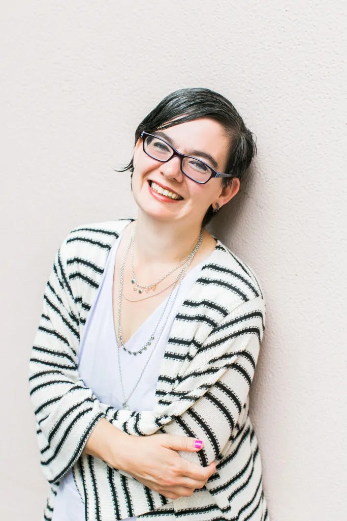 Cindy, a white woman with short dark hair and glasses, smiles at the camera. She is leaning against a light-colored wall and wearing a striped cardigan, light top and several necklaces