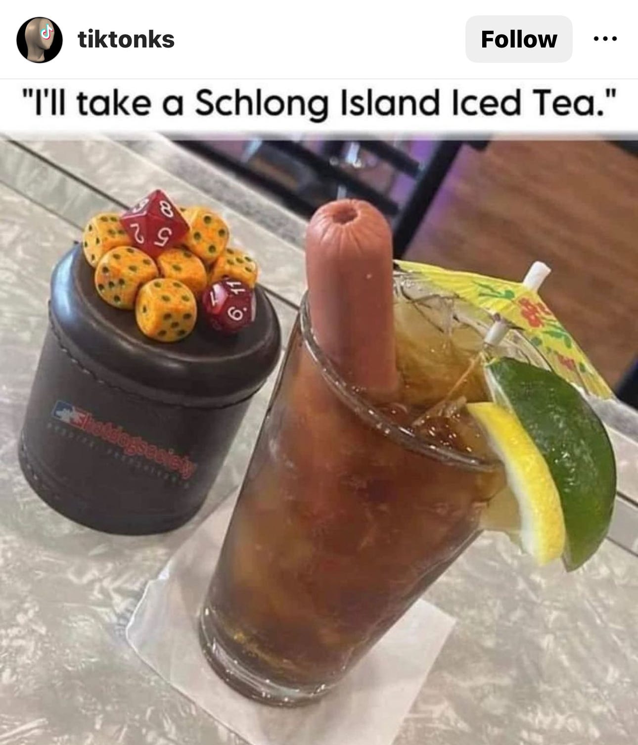 A drink that looks like a Long Island Iced Tea with a slice of lemon and lime on the rim and a cocktail umbrella and hot dog in the drink. Captioned: "I'll take a Schlong Island iced tea."