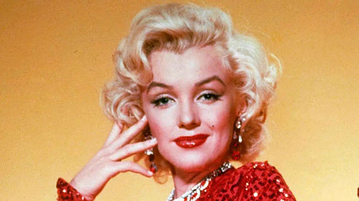 Image description: Marilyn Monroe's face and shoulder is pictured, she has her hand held to her face. She is wearing a red sequined dress, red lipstick, and has a subtle smirk on her face.