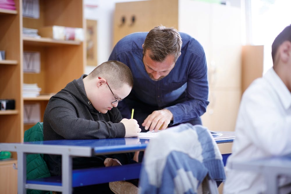 Classroom. Male teacher helping boy with Down's Syndrome with his written work