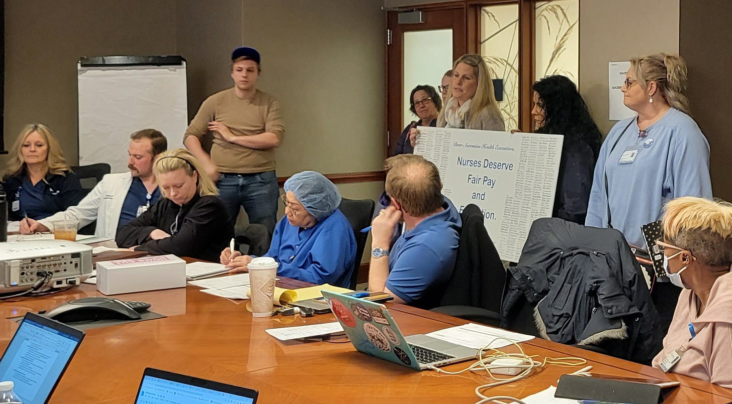 a group of people wearing scrubs and sweaters standing and sitting around a table in an office. a blank white poster board is to the left of the image and a poster board reading "nurses deserve fair pay" with tiny text stands in front of people holding it up