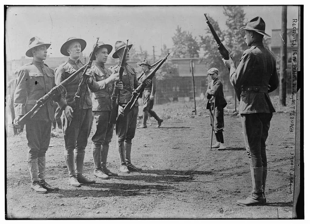Black and white photo from 1915 depicting soldiers conducting drill at recruit training. Four soldiers are in the left front foreground with rifles receiving instruction from a soldier standing in front of them. That soldier is at attention and holding his rifle at port-arms.  In the background is one other soldier marching and another informally observing those receiving instruction. 