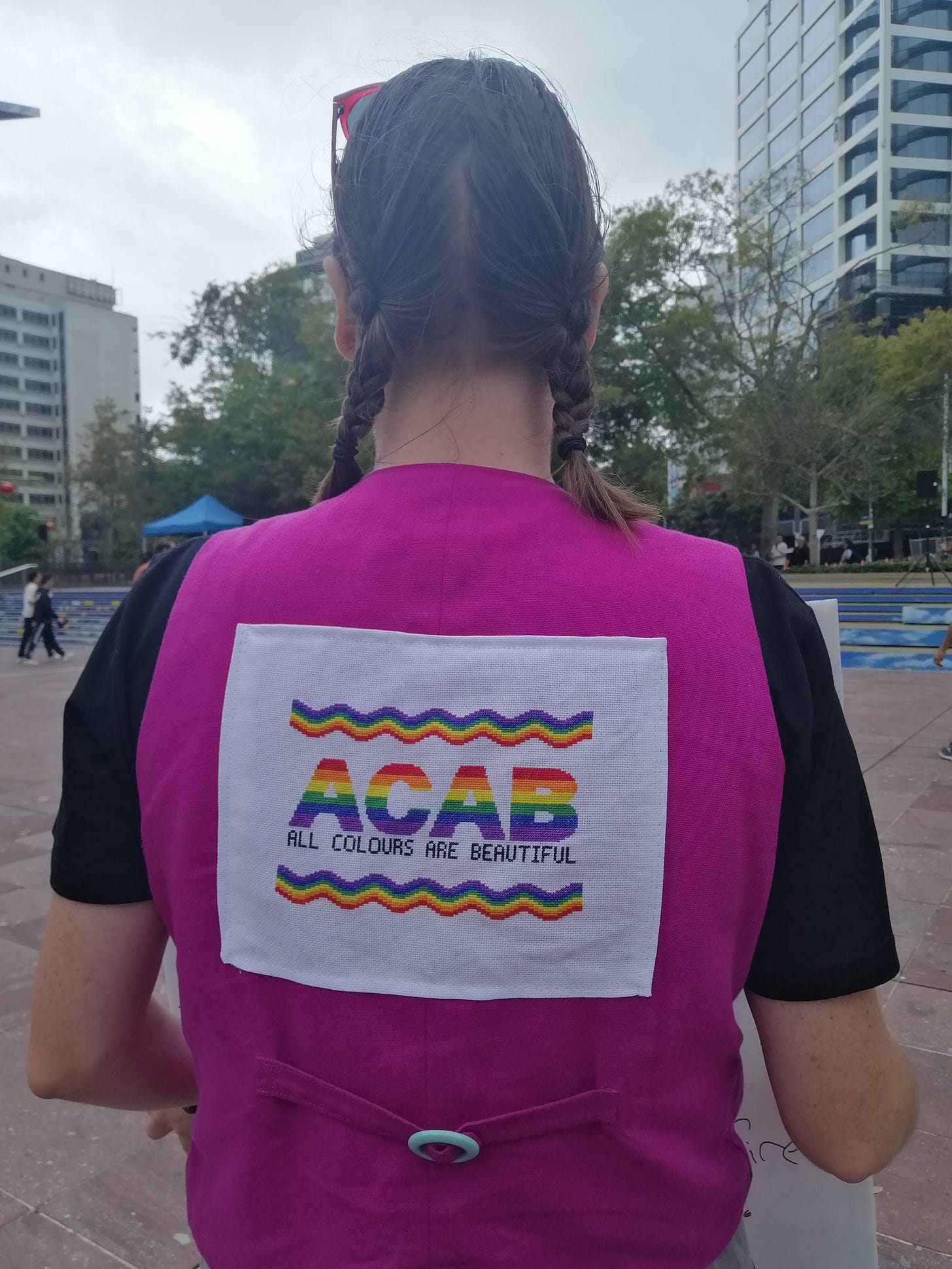 Photo of a white woman with shoulder length brown hair in two plaits wearing a hot pink vest with ACAB stitch on the back