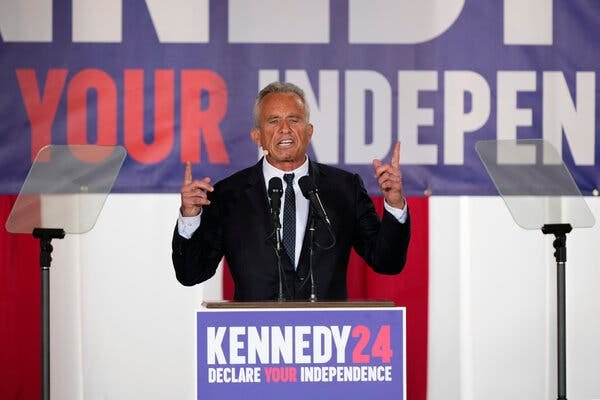 Robert F. Kennedy Jr., wearing a black suit, points upward while speaking on a stage with signs that read “Kennedy 24 Declare your independence.”
