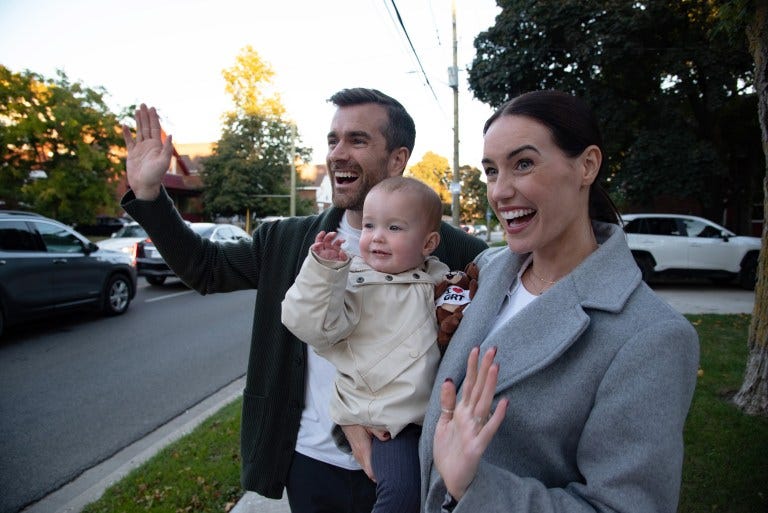 A smiling man on the left and a smiling woman on the right holding a baby between them. All three are waving.