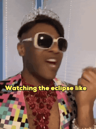 giphy of Black man with short. hair, sunglasses, a tiara, red jeweled necklace and multicolered low cut shirt mouth "oh my god" and move his hands in emphasized mannerisms with yellow text in the middle that says "watching the eclipse like"