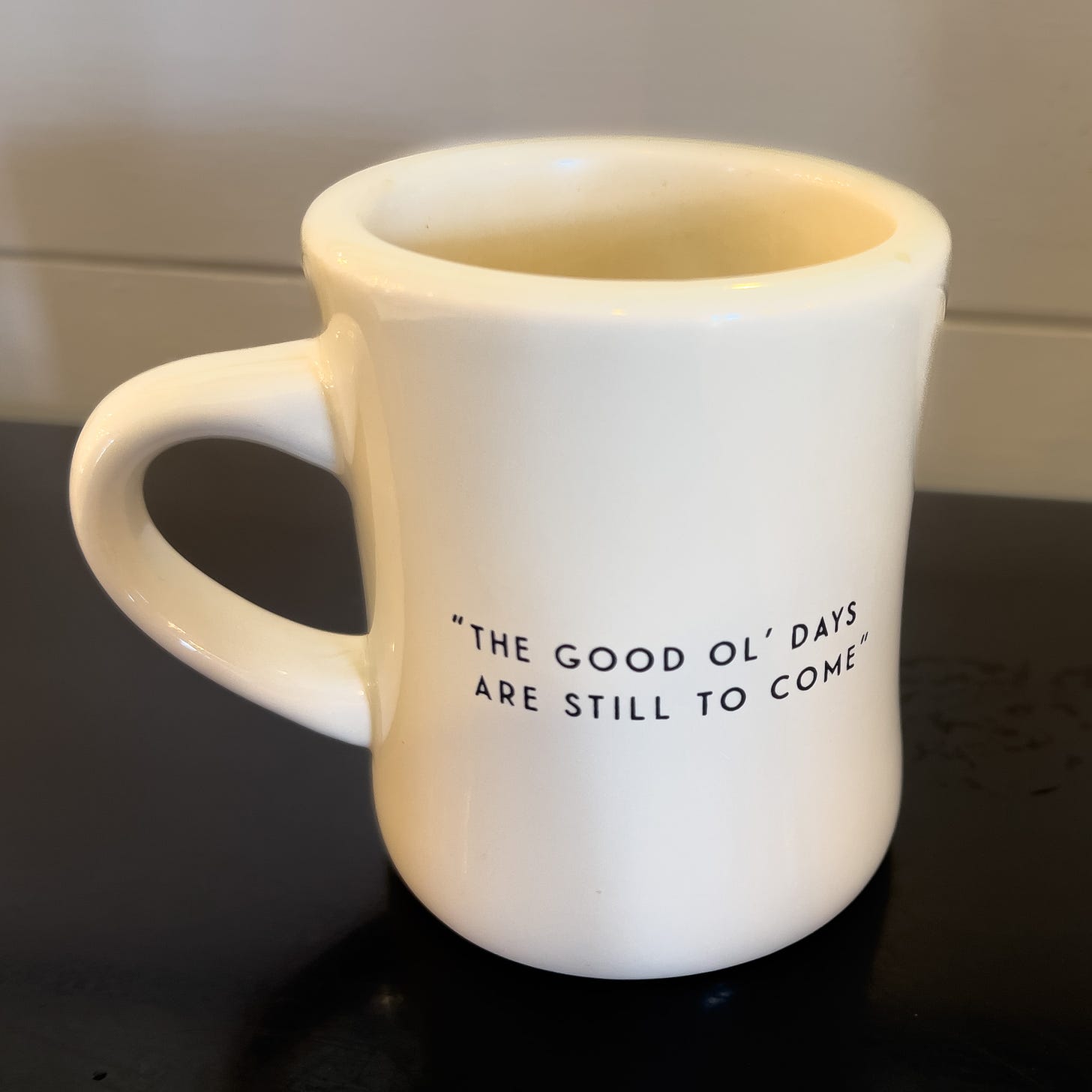 A white mug sitting on a balck table with the words” “The good ol’ days are still to come.”