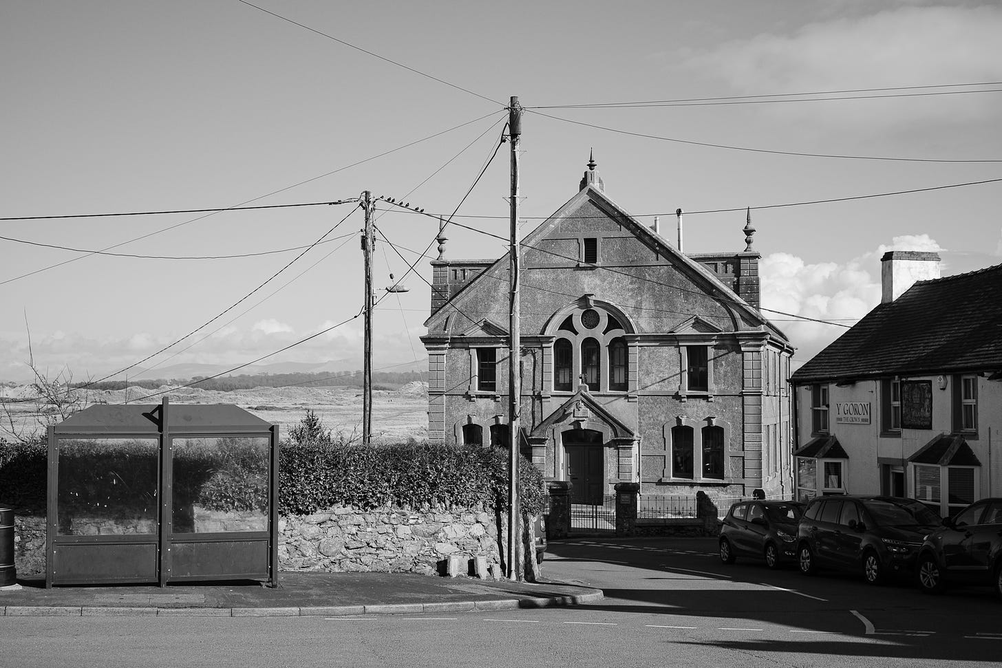 Black and white photograph of a street scene featuring a bus shelter, telegraph pole and wires in front of a former chapel and a pub on the right called Y Goron