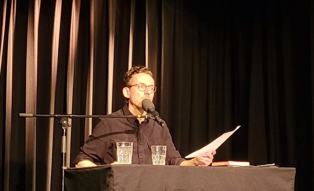Auf einer dunklen Bühne sitzt Paul Bokowsi an einem schwarzen Tisch. Er trägt ein schwarzes Hemd, hält ein Blatt Papier in der Hand, schaut zur Seite und spricht ins Mikrofon. 