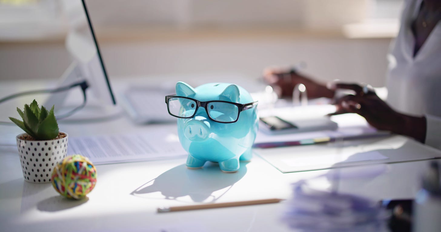 A big blue piggy bank wearing a pair of glasses as his human companion works nearby on a laptop