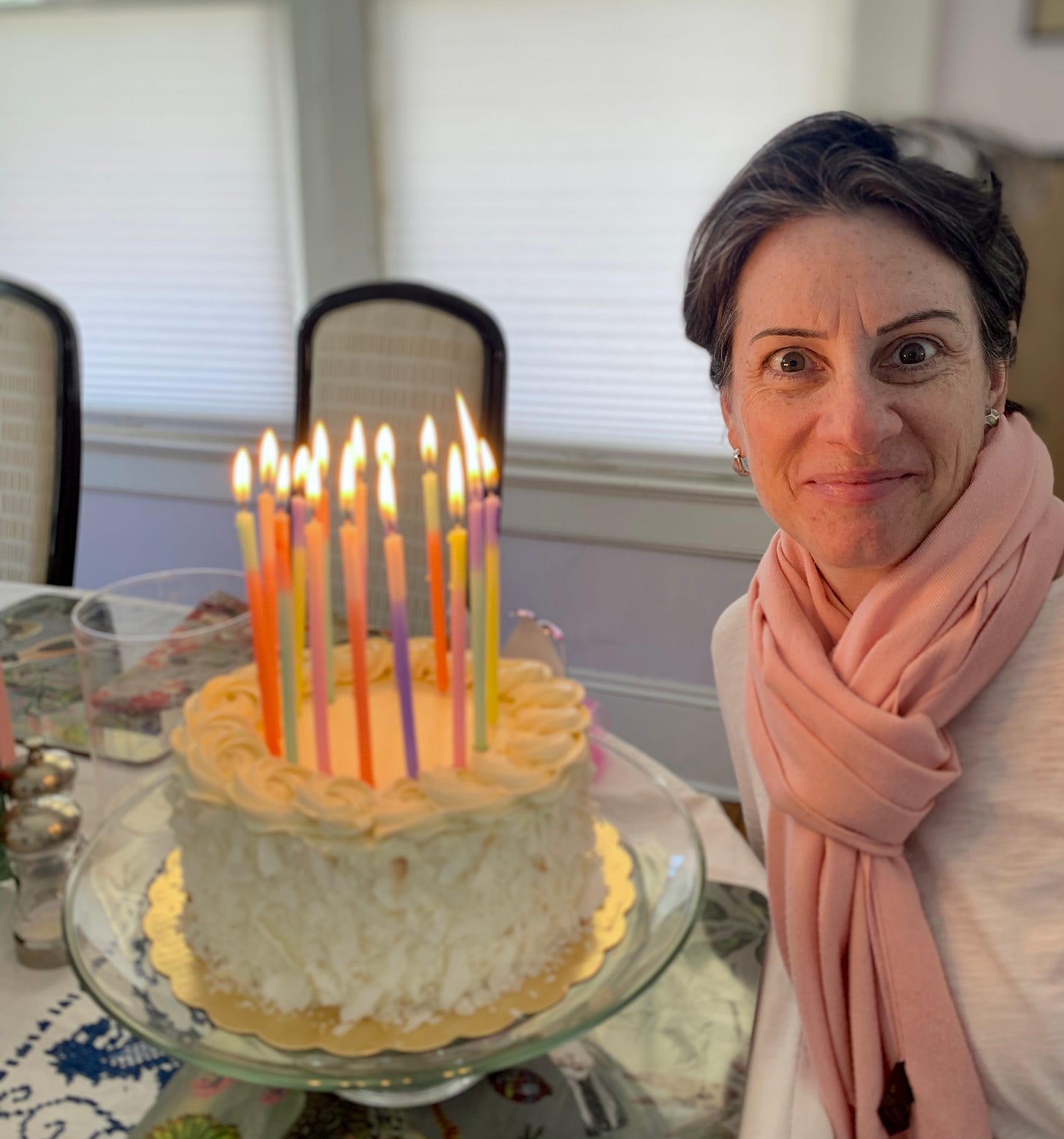 Elizabeth smiling, wearing a pink scarf, in her dining room with a lemon cake and plenty of birthday candles in front of her last year.