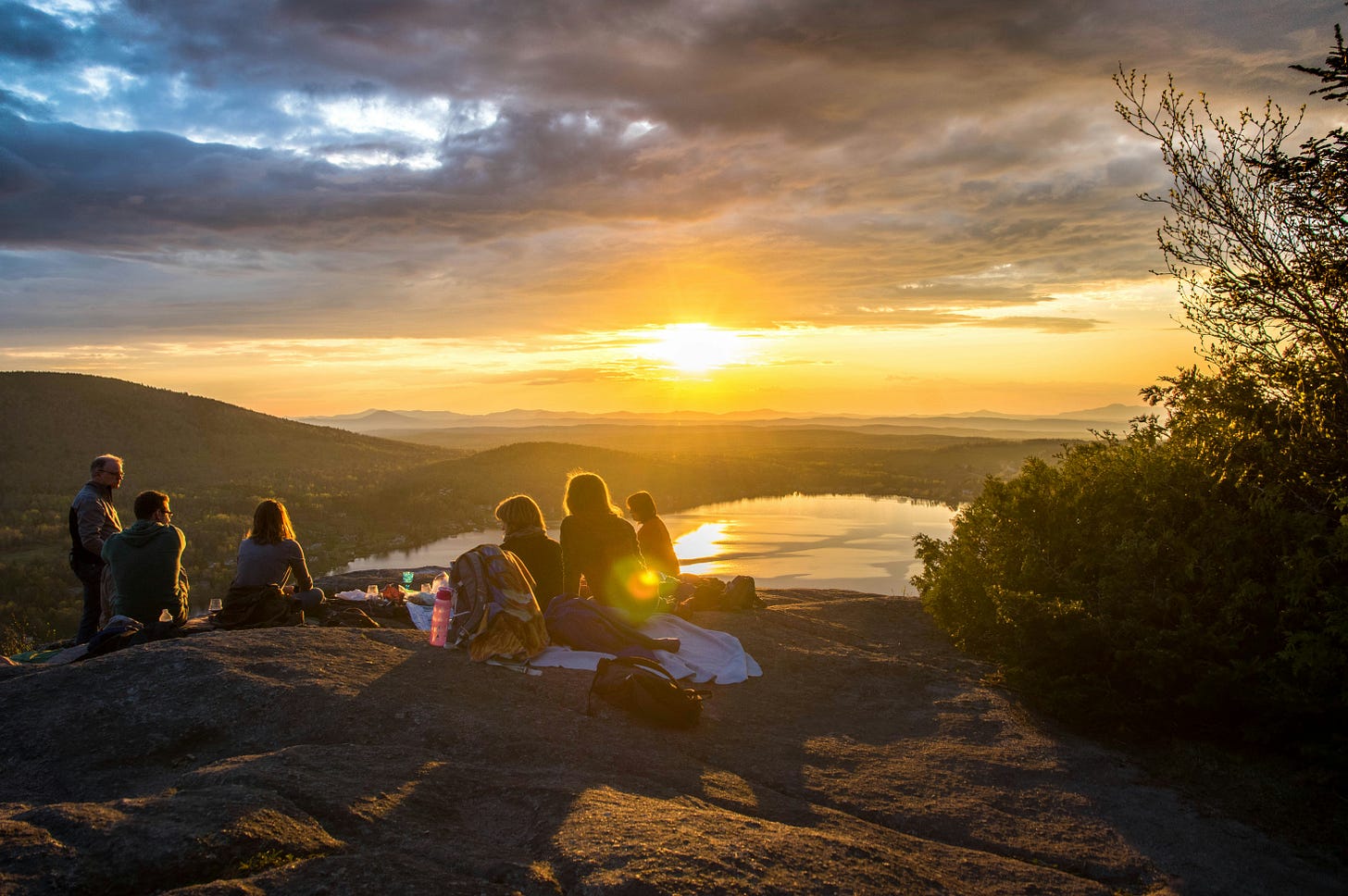 Image of people on a cliff overlooking a mountain lake, enjoying a sunset picnic.