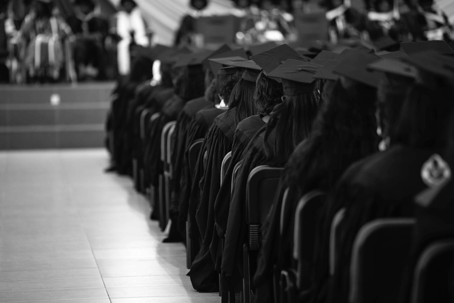 A black and white photo of graduates sitting in audience. They are seated one in front of another. The aisle is on the left.