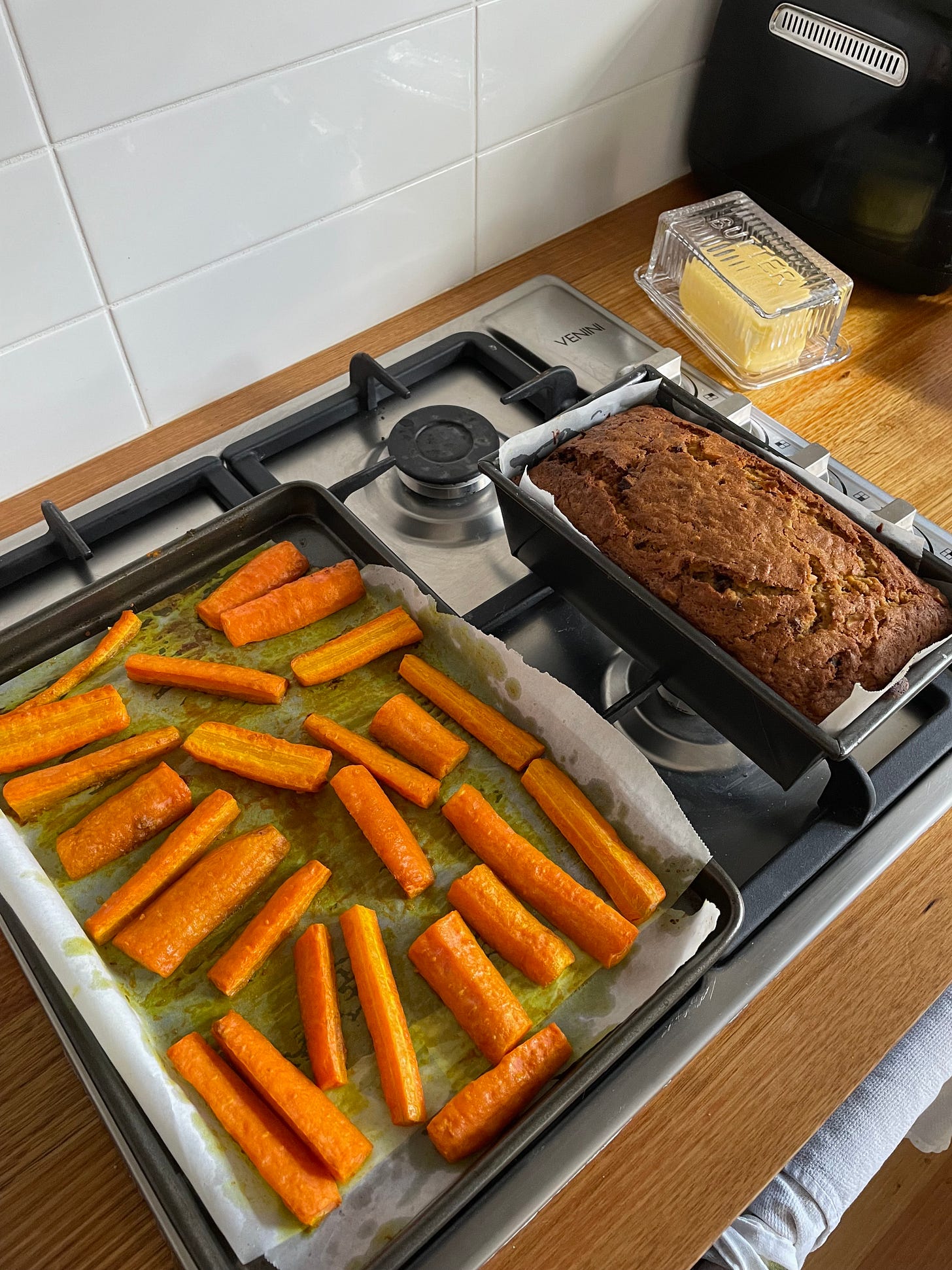 Tray of roasted carrots on a stove next to a just-baked carrot cake loaf