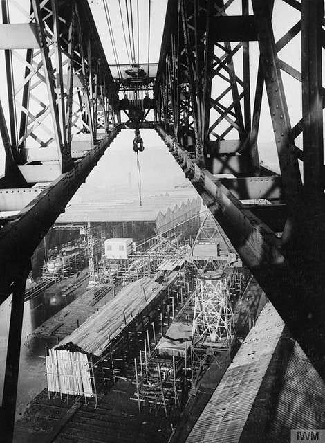 A black and white photo, taken from the top of a crane, looking down on a few sheds and scaffolding structures. Apparently it’s a shipbuilder’s yard, but I don’t know how they can tell as the only clue is the sea in the distance.