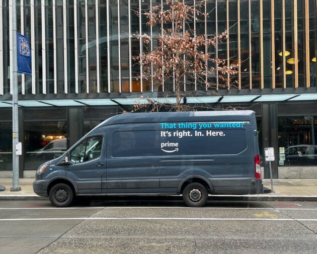 Image shows a blue Amazon delivery van parked in front of an office building. The side of the van says, "That thing you wanted? It's right. In. Here."
