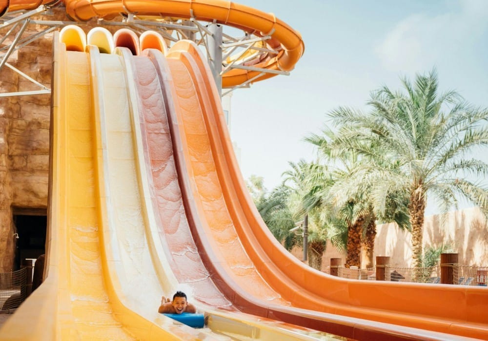 Child on blue inner tube going down orange and yellow twirly slide by palm trees