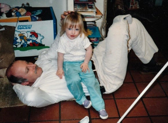 the author as a tiny child sits on her dad's torso as he is lying down on the floor in a kind of sit-up position with one hand behind his head and one hand ruffling his daughter's hair. he is smiling and looking at her while she looks at the camera