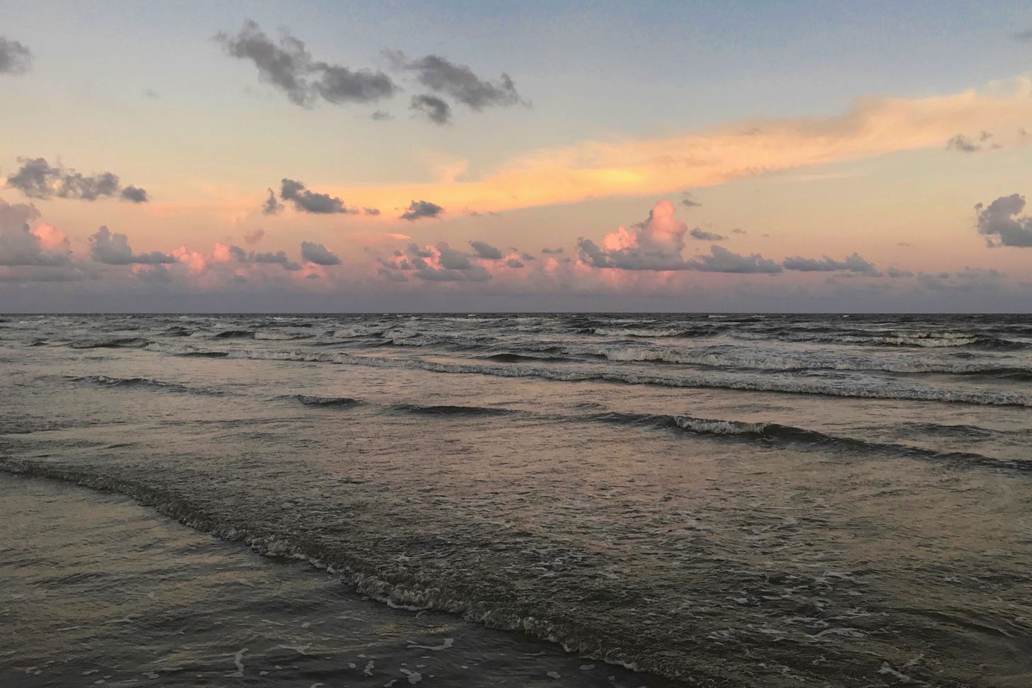 Waves roll onto the beach at sunset with clouds lined with corals, pinks, and grey and reflected in the water