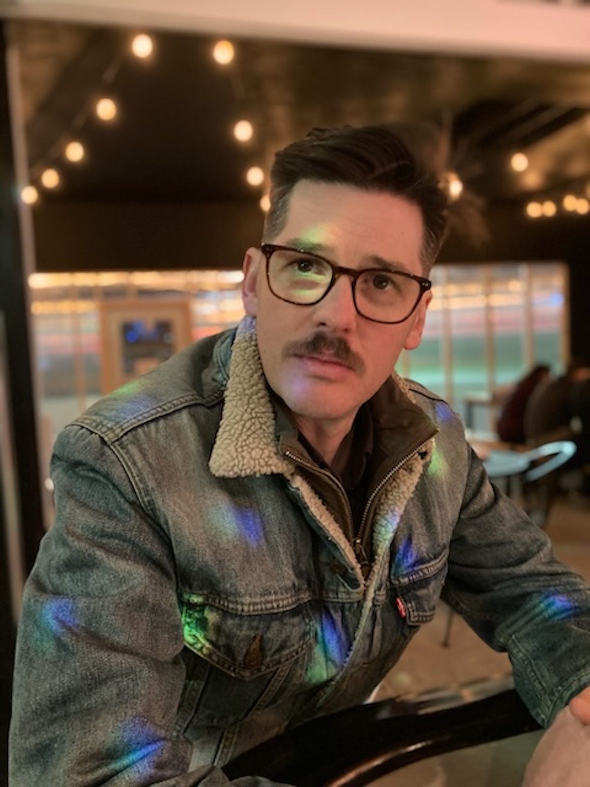 Author Andrew Ewell looks at the camera while sitting in a restaurant. Ewell has a tight, cropped haircut, a pair of black glasses and a mustache. He's wearing a jean jacket. 