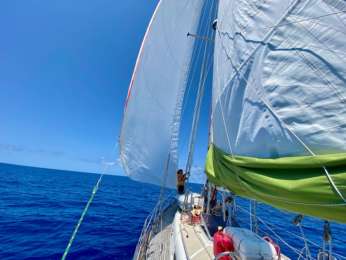 Image: A brilliant blue sky over a brilliant blue ocean with a sailboat in the foreground as seen from the stern looking forward. 