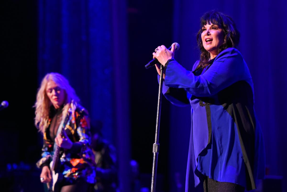 A blond woman playing a guitar stands on stage with a dark-haired woman who holds on to a mic and its stand