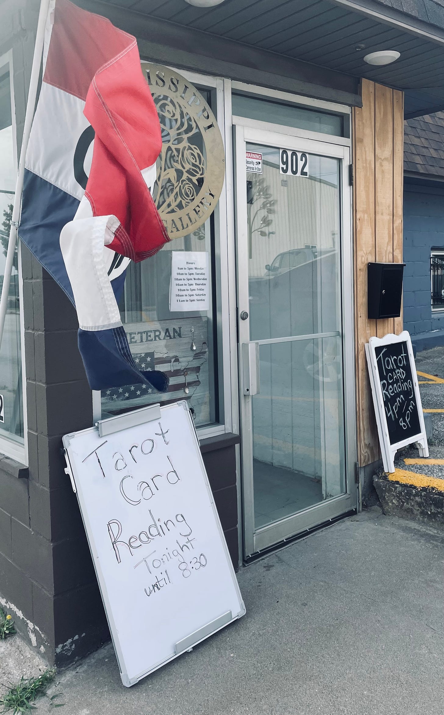 A red, white and blue flag with the word "Open" waves in the wind outside a business door, where there are also two signs advertising tarot card readings happening inside.