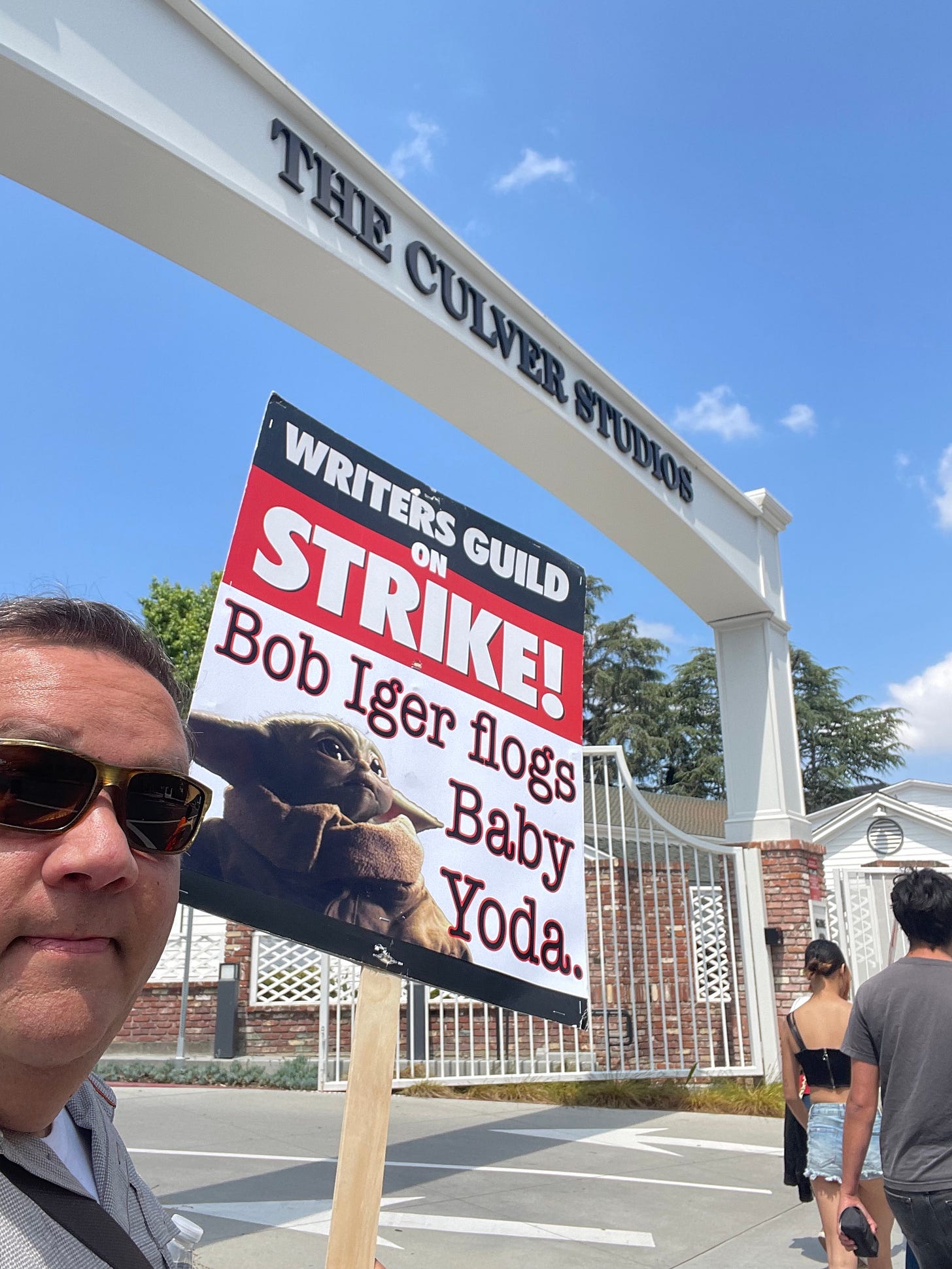 A picture of Mickey holding a sign that says "Bob Iger flogs Baby Yoda" with a picture of Grogu, Mickey is standing underneath the main gate at Culver Studios.