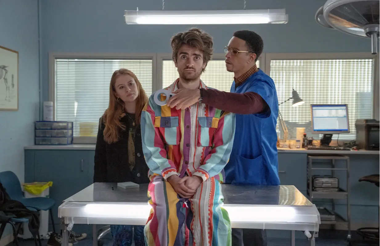 Actors Máiréad Tyers, a white woman with long red hair wearing a dark jacket, and John MacMillan, a Black man with glasses, short curly black hair, and a goatee, wearing blue scrubs over a long-sleeved maroon top, stand behind a metal table in a veterinary office. Between them, on the table, sits actor Luke Rollason, a white man with medium-length wavy brown hair and facial hair wearing brightly colored pajamas, as MacMillan passes a round white medical scanner over his chest in the Disney+ series Extraordinary.