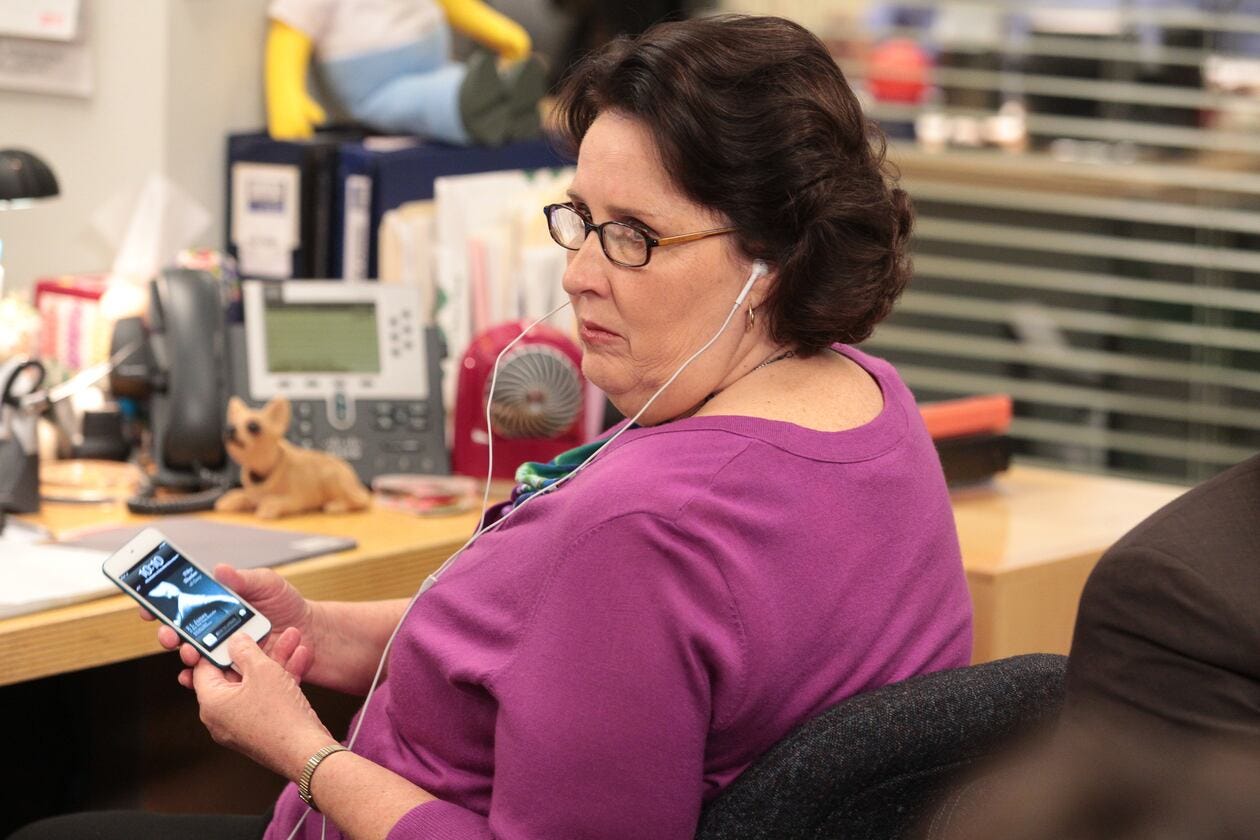 cena de The Office com a personagem Phyllis: uma mulher branca de cabelos castanhos, óculos de grau e blusa rosa está sentada numa mesa de escritório ouvindo com fone de ouvido o audiolivro Cinquenta tons de cinza