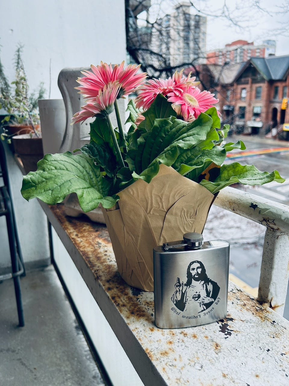 On a balcony, a potted plant of mums beside a flask with an image of Jesus on it and the words "What would Jesus do?"