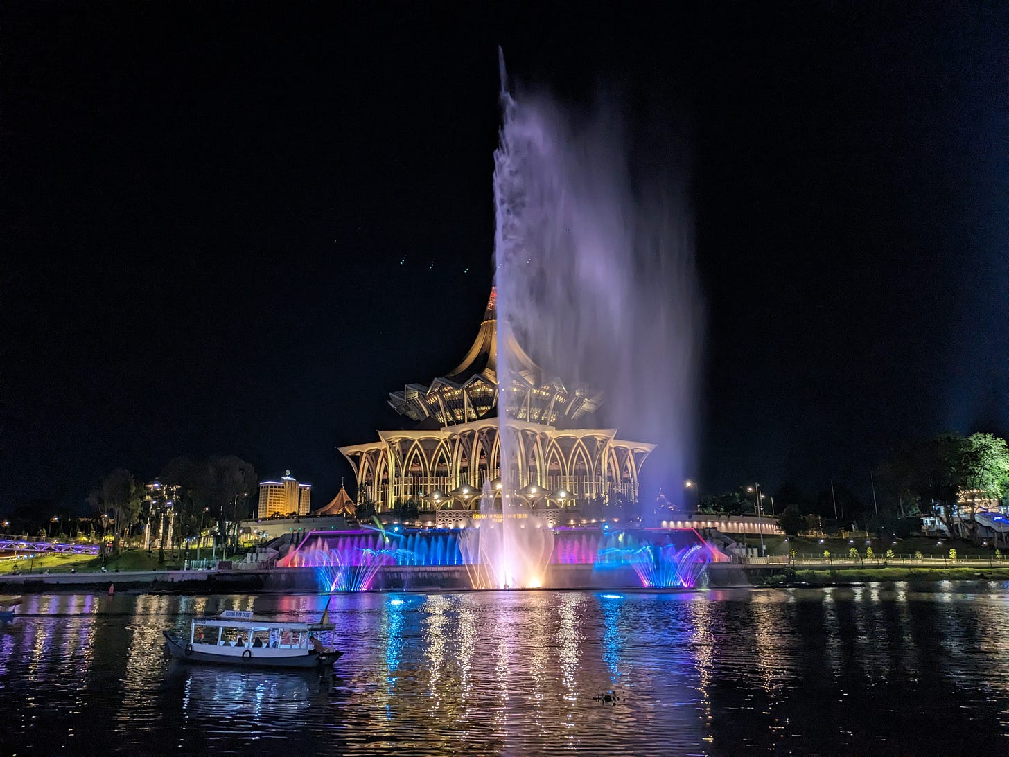 a fancy and important, exotic-looking building lit up at night by lasers and colorfully lit fountains, across the peaceful river with a small boat in the foreground, carrying passengers who are watching the show