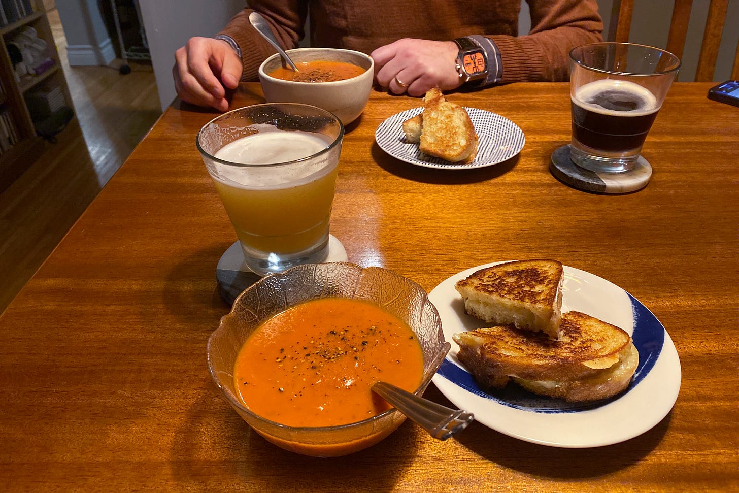 A spread of tomato soup in small bowls and grilled cheese on small plates. The soup is topped with cracked pepper and the sandwiches are cut in half. Glasses of beer are on coasters between the two dinners, and Jeff's hands are on the table near his bowl of soup.