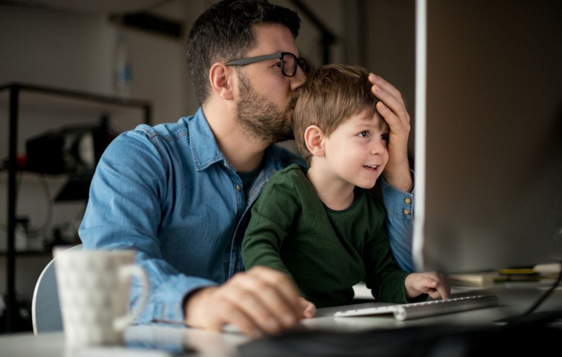 A working dad with his kid on the lap giving the little boy a kiss, while he glances over to the laptop screen