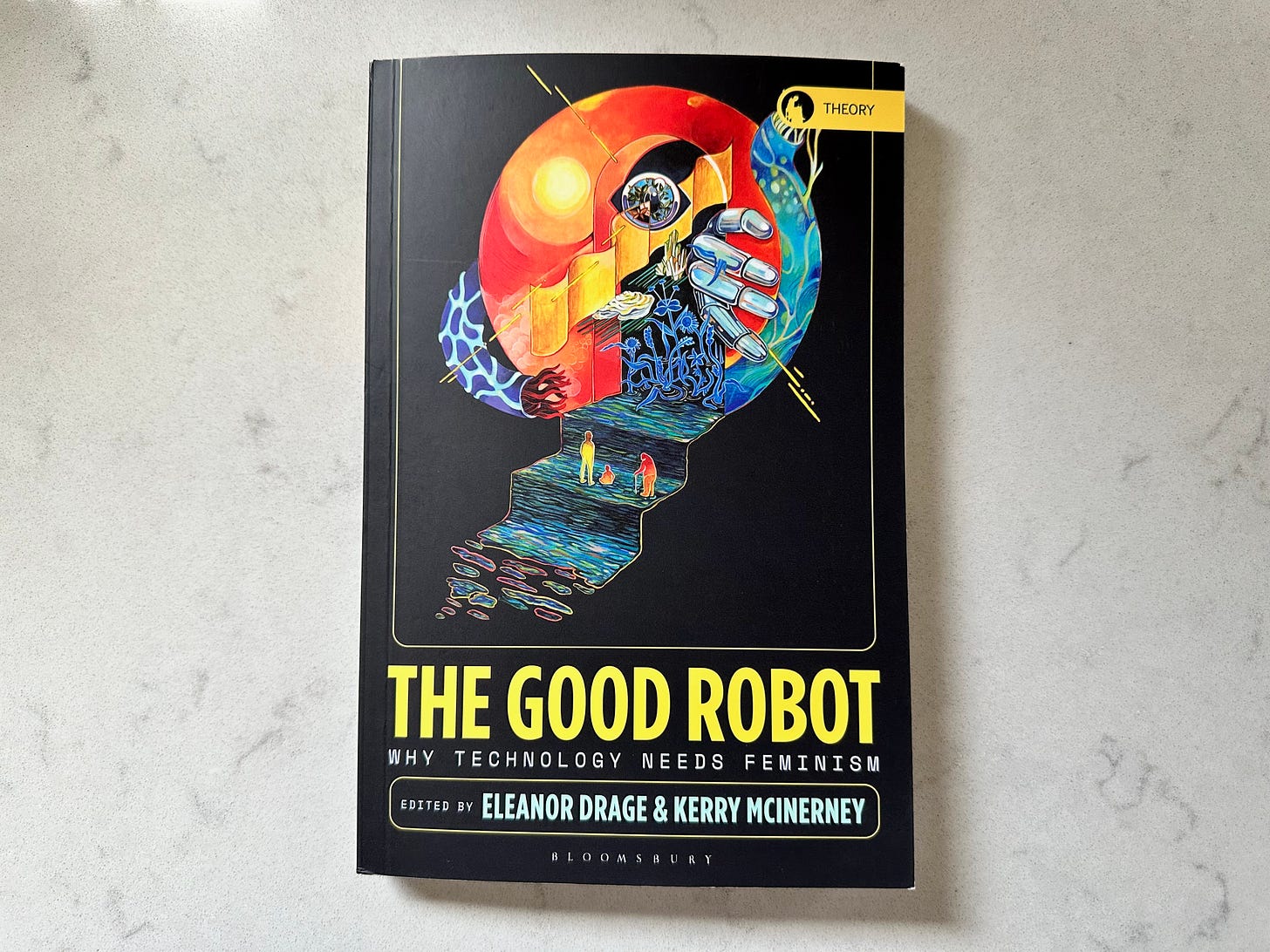 Picture of a book titled "The Good Robot", edited by Eleanor Drage and Kerry McInerney, on a white marble table. The cover has a futuristic, abstract artwork by illustrator Sinjin Li showing three figures climbing a styled staircase towards fiery red and blue sphere.