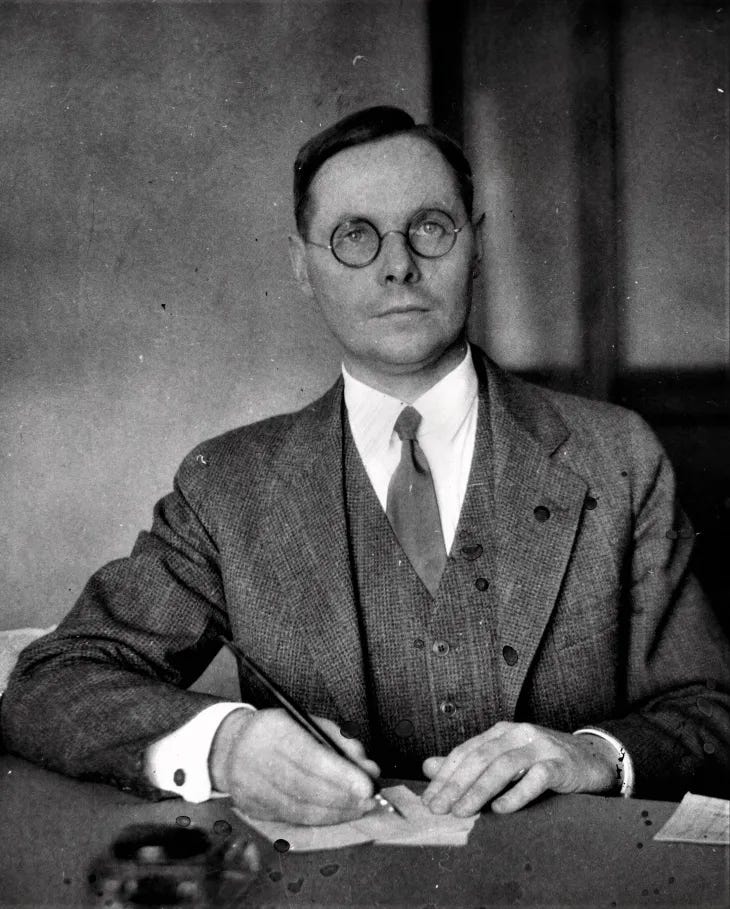 a black and white grainy photogrpah of a white man with round glasses looking unsmilingly at the camera. He is wearing a three piece suit and a tie