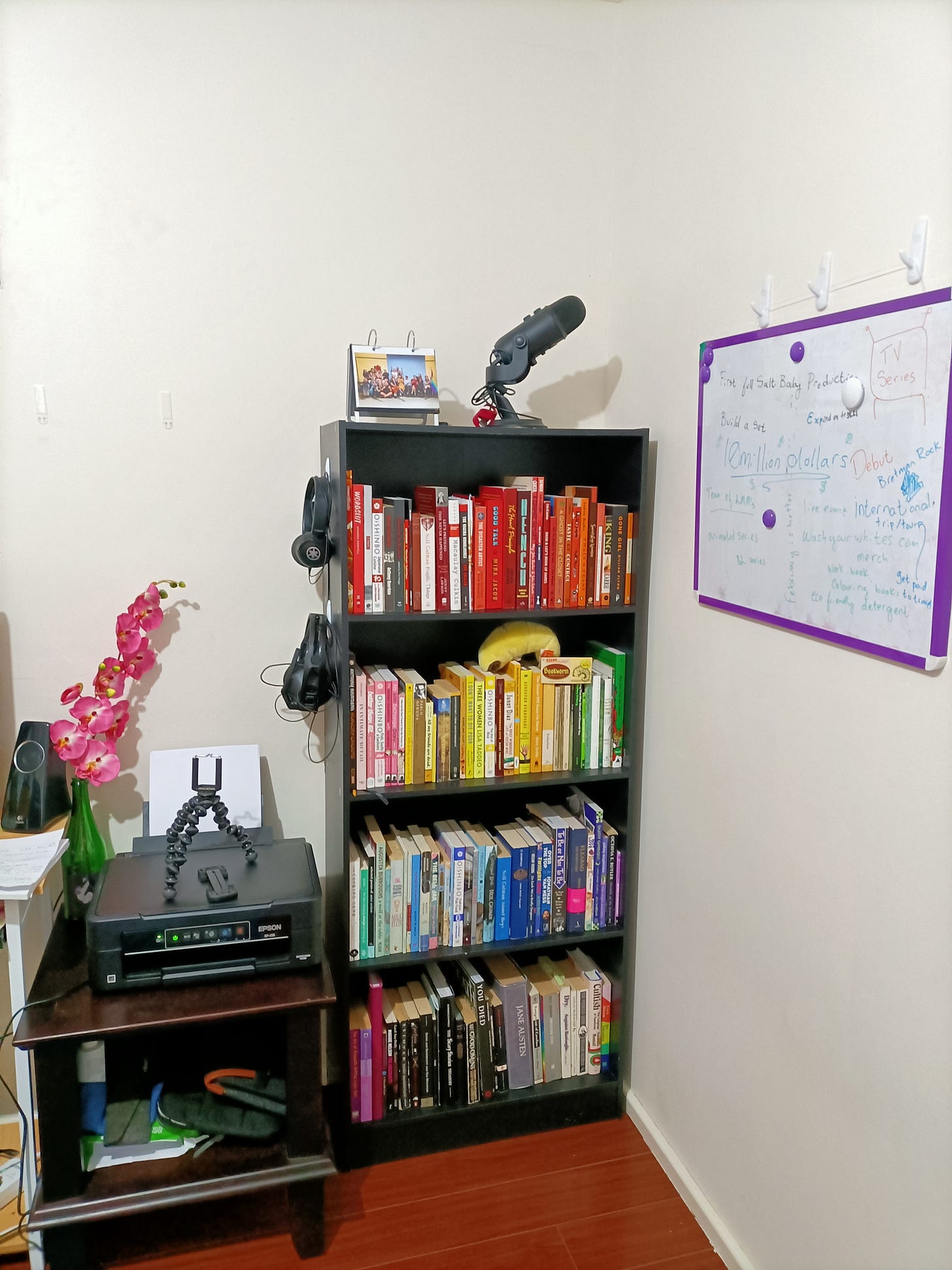 A photo from Miranda's home office. On the left, you can see the printer on a side table, next to a green bottle with a fake orchid. To the wall on the right, you can see a hanging whiteboard scribbled with dreams. Centre of the photo is their pride and joy, their rainbow organised bookshelf. It goes from red on top to black/white/grey on the bottom, and includes a variety of fiction, non-fiction, graphic novels and more.