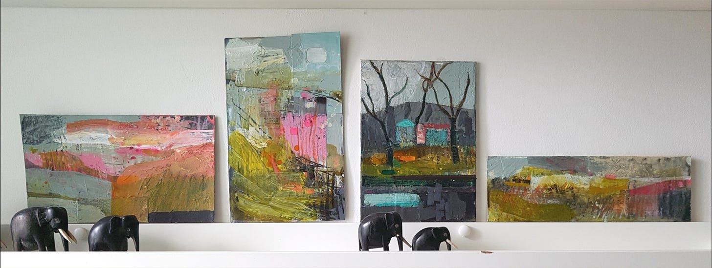 Small colourful abstract landscape paintings on a white shelf against a white wall. There are several carved elephant ornaments on the shelves beside the paintings.