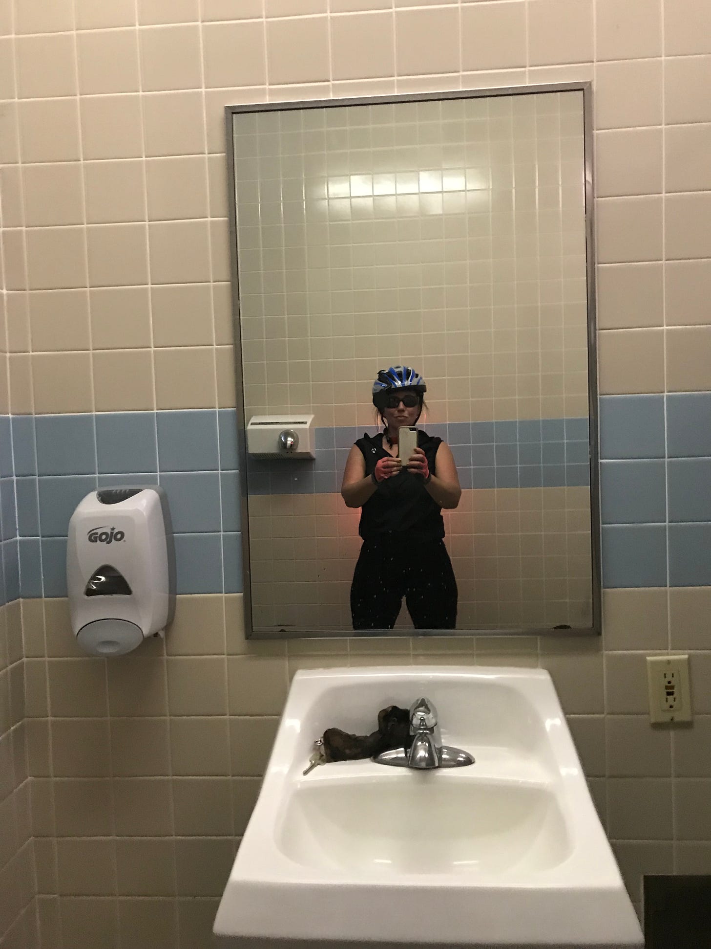 A woman wearing a helmet and bicycling gloves and gear taking a selfie in a bathroom mirror