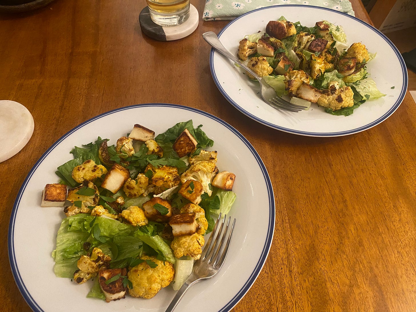 The salad described above, on white plates with blue rims and forks resting at the edges: green lettuce mixed with charred roasted cauliflower, crispy cubes of halloumi, and bits of parsley throughout.