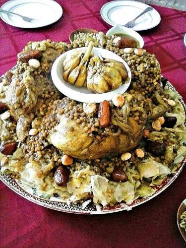 A platter of a Moroccan chicken and lentil stew served over a bed of shredded pastry. A whole chicken is centered on the pastry bed. Whole garlic cloves sit in a bowl on top of the chicken. The entire dish is garnished with dates and fried almonds.