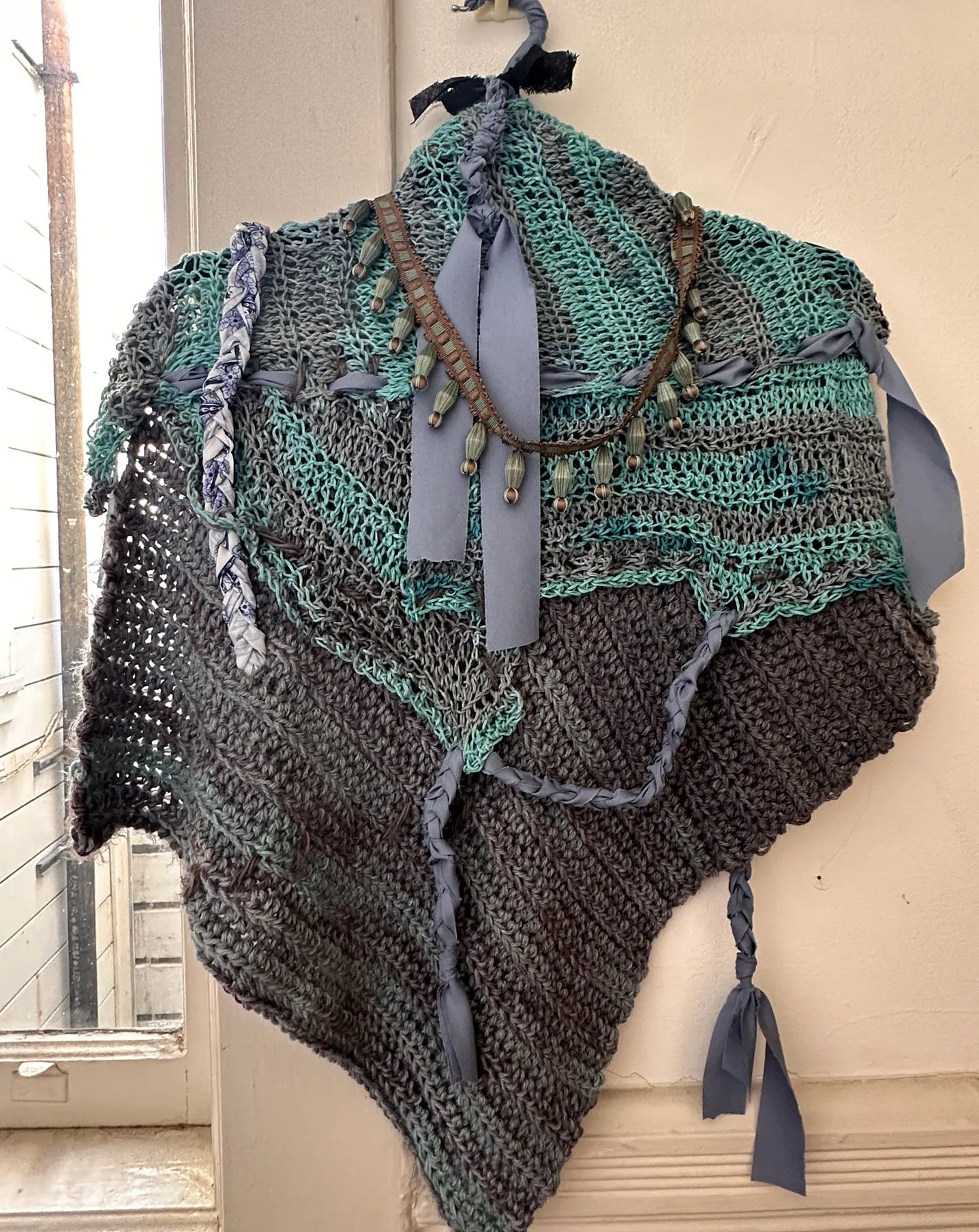 A teal and gray crocheted scarf on a hanger, with a beaded green necklace draped across the front