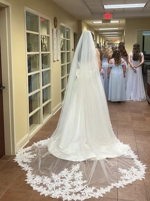 Rearview of the bride with epic cathedral veil spread big, bridesmaids ahead of her in the hall waiting to turn and look