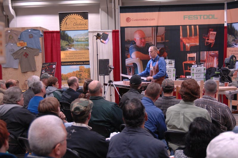 Andy teaching a seminar from a platform full of tools and furniture, surrounded by a large crowd