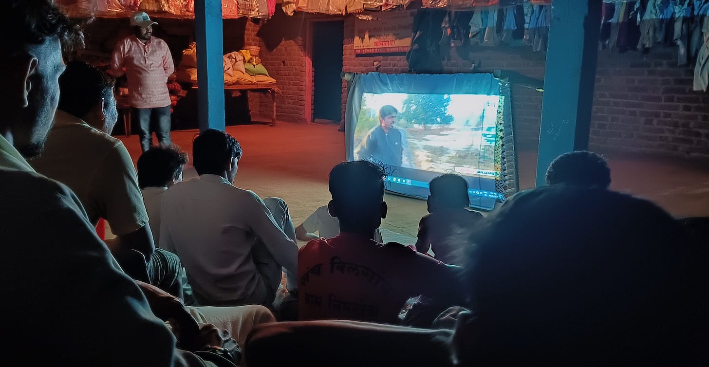 A group of people sitting in a dark room watching a short video being screened on a white screen attached to a charpai A group of people sitting in a dark room watching a short video being screened on a white screen attached to a charpai