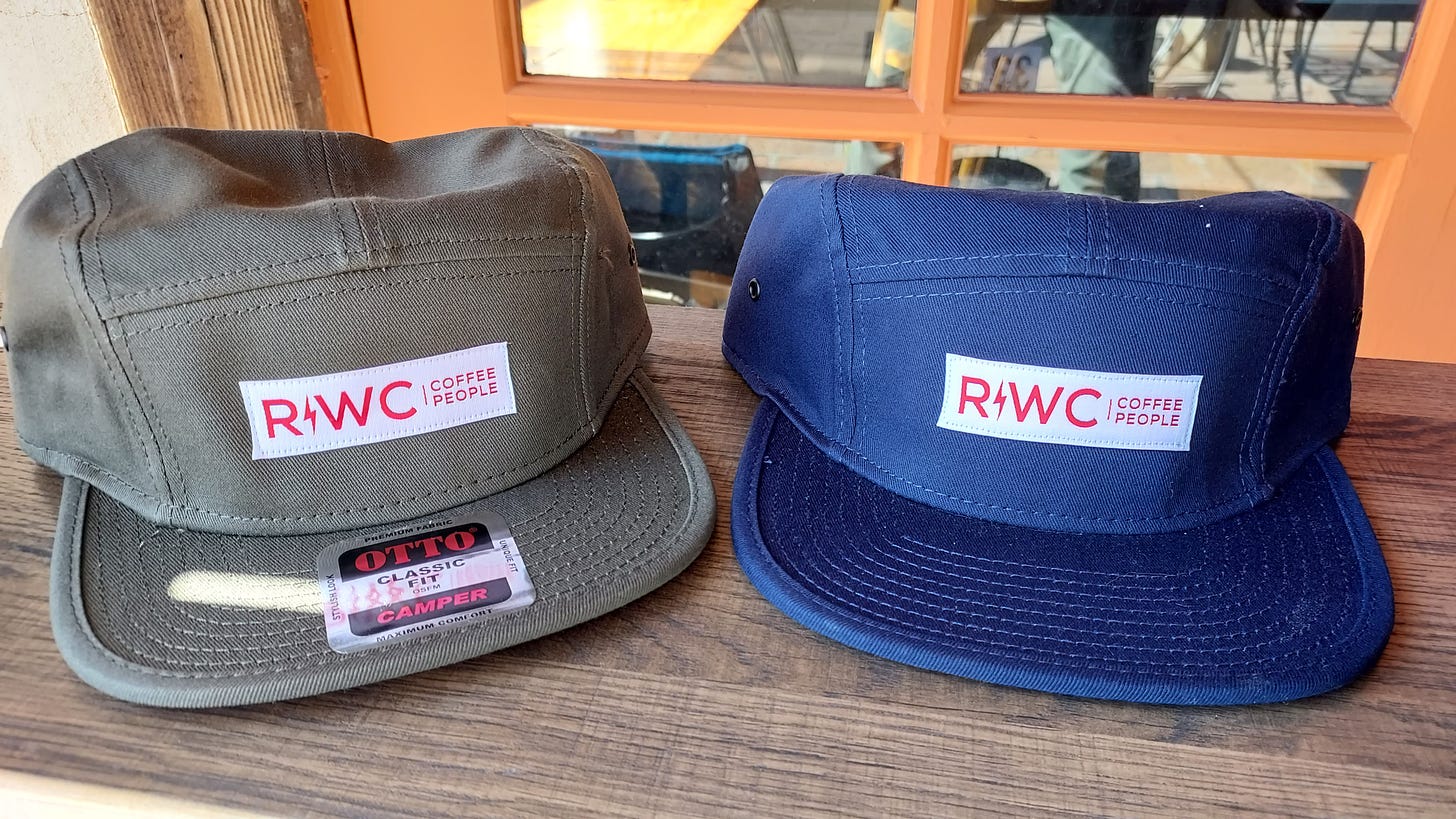 From Left: A grey 5-panel camp style cap with the R!WC Coffee People Logo and the same in nave sit on a wooden shelf.